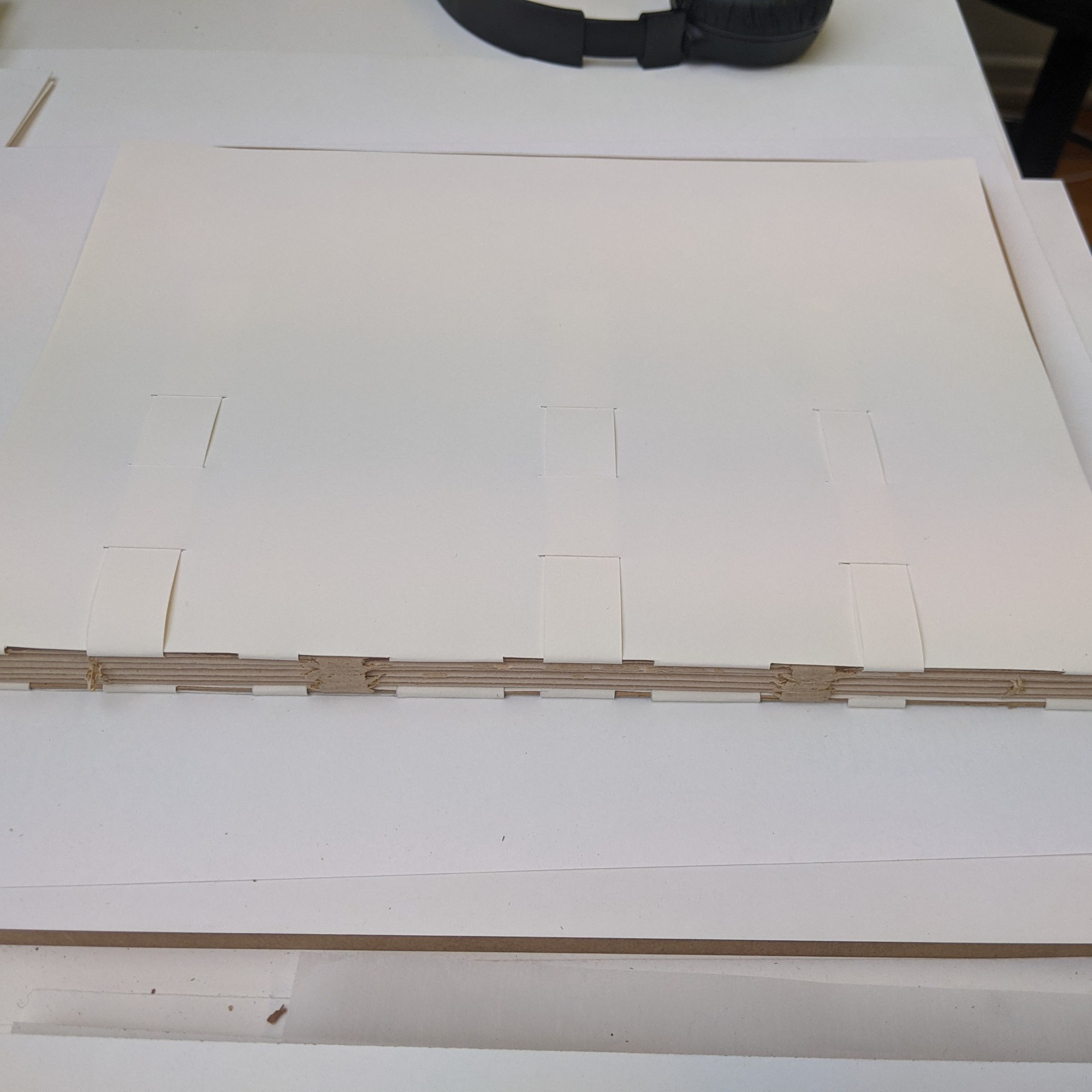Image shows the construction of a wrapper which wraps around the board of the book around the spine edge. It is made up of two sheets of buffered archival paper which has tabs cut out that pass around the spine edge of the board to the other side of the board. Then the tabs are woven into slots in the corresponding wrapper.
